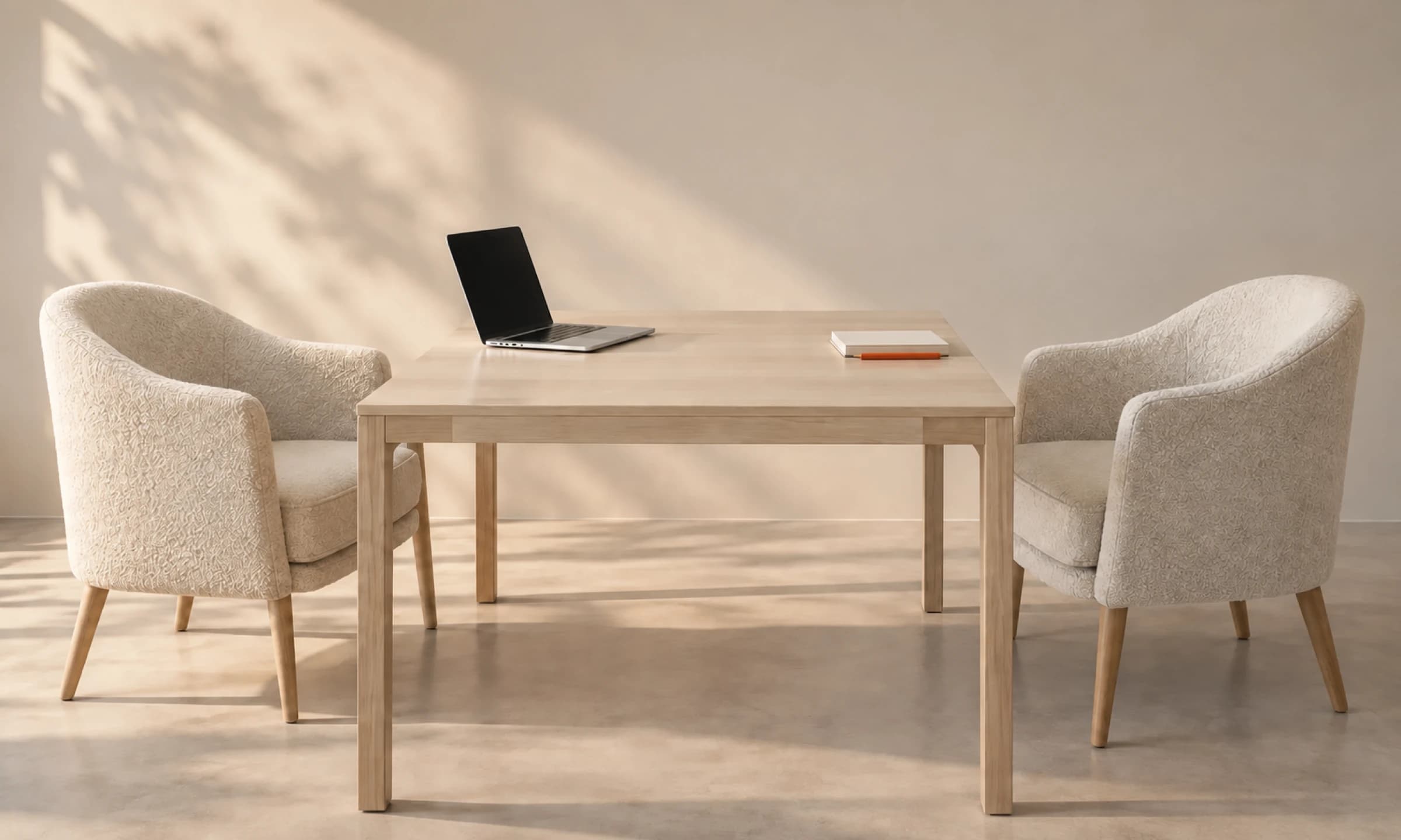 Zwei cremefarbene Sessel stehen sich an einem hellen Tisch in einem minimalistischen Raum gegenüber; auf dem Tisch ein Laptop, ein Notizbuch und ein orangefarbener Stift.