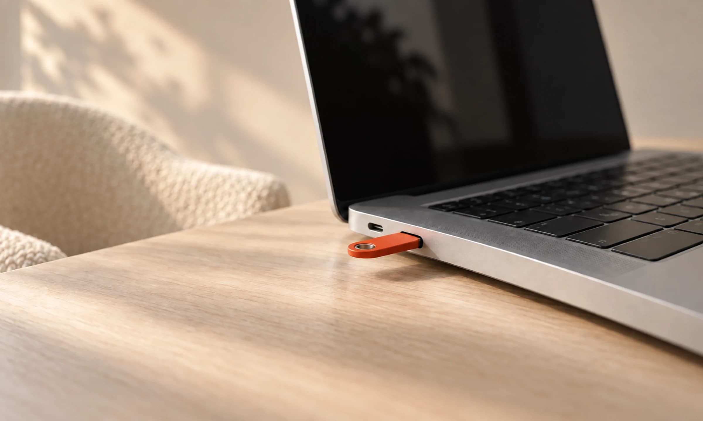 Ein orangefarbener Sicherheitsschlüssel steckt seitlich in einem geöffneten Laptop; im Hintergrund ein cremefarbener Sessel.